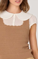 A woman is wearing a white short-sleeved blouse with a rounded collar that has scalloped edges, layered underneath a sleeveless, brown crochet-knit top.